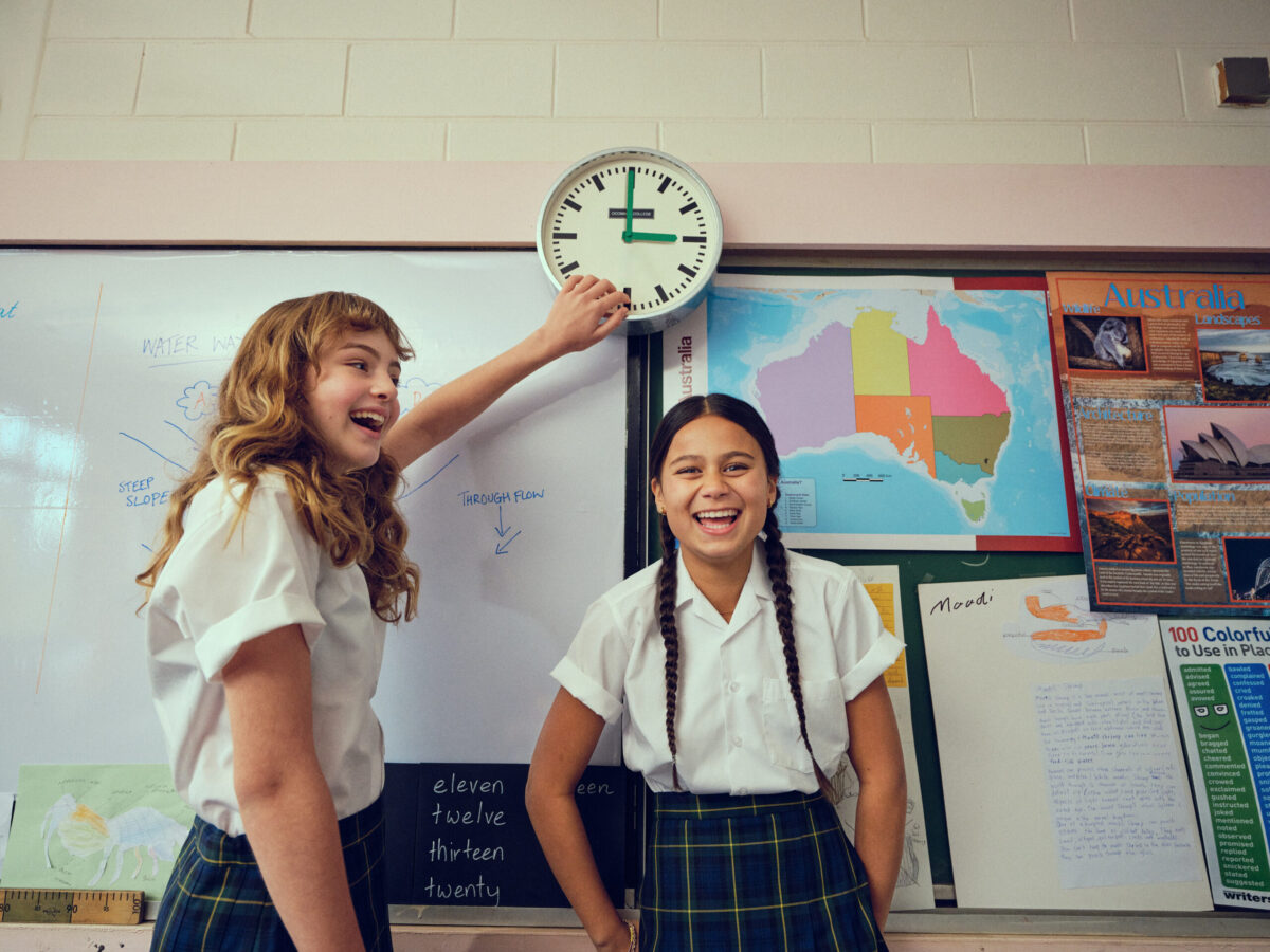 An image of school girls in a classroom pointing at a clock
